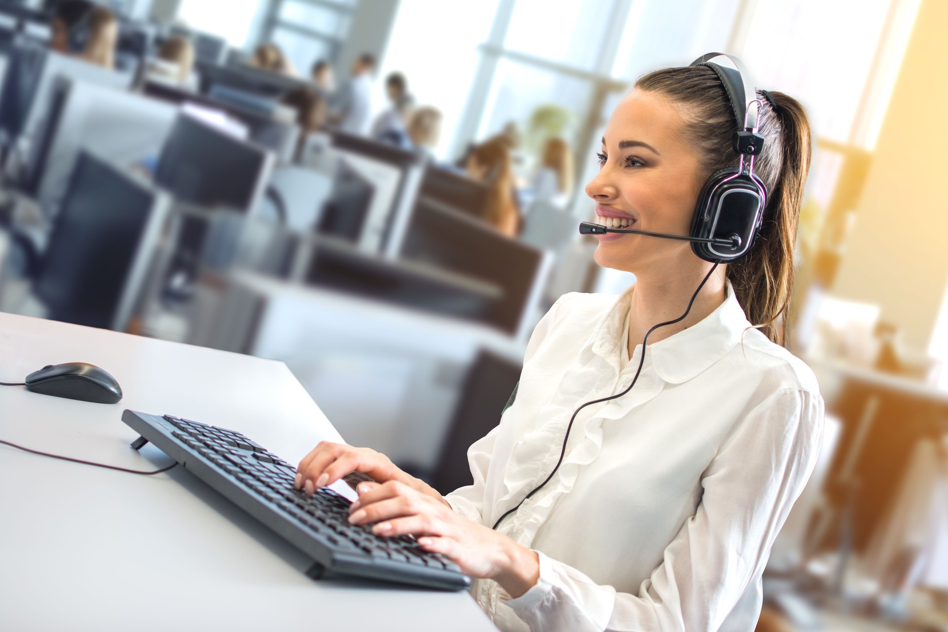 Friendly female helpline operator working on her workspace at large busy call center office.